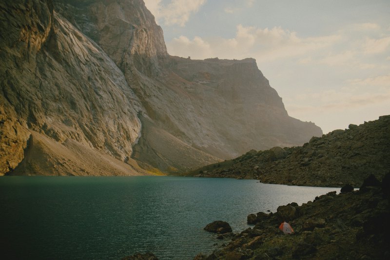 A small orange tent on a rocky shore beside a turquoise mountain lake